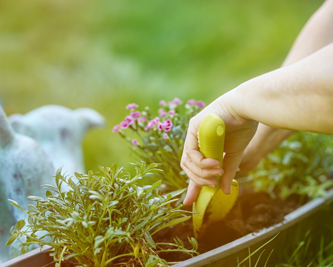 Hands carefully plant vibrant small flowers into a rectangular planter box with a yellow trowel, surrounded by lush greenery and soft natural light, highlighting spring gardening.