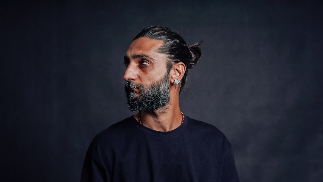 A high-resolution portrait of a bearded man wearing a casual t-shirt and a single earring, set against a dark background. Subtle, moody lighting emphasizes his facial hair, accessories, and modern masculine style — ideal for editorial, lifestyle, and fashion projects.