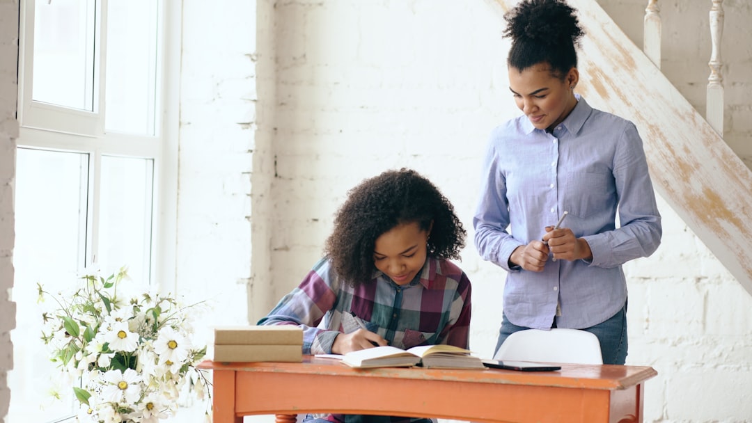 Teenage curly haired mixed race young girl sitting at the table concentrating focused learning lessons and her elder sister helps her studying at home